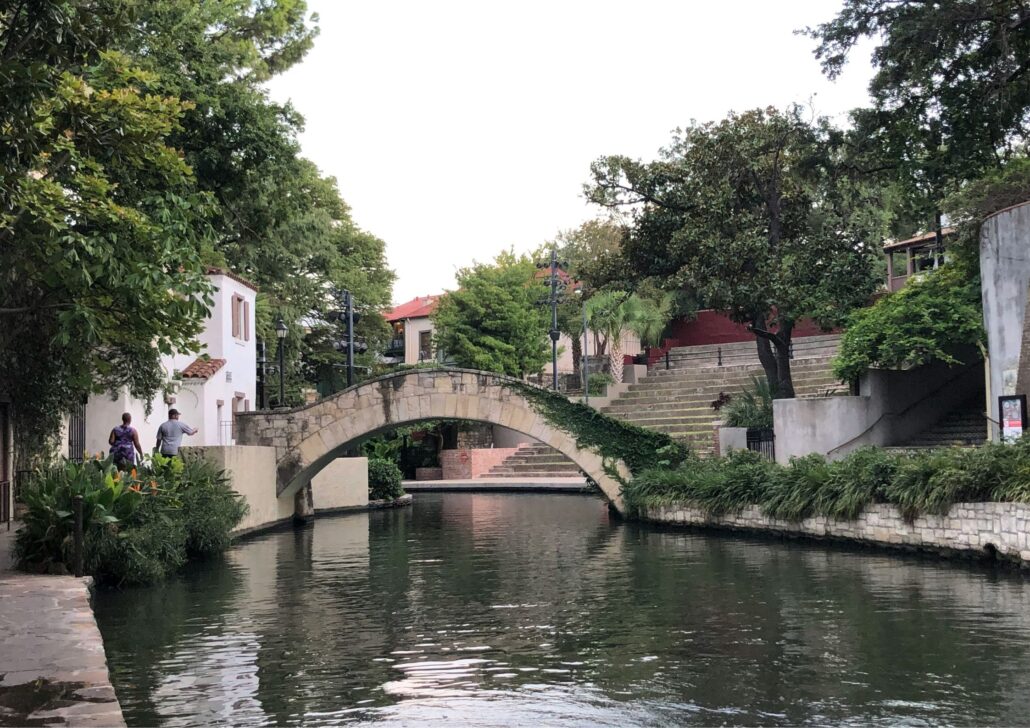 Scenic view of a stone bridge over a calm waterway in San Antonio, Texas, surrounded by lush greenery and residential buildings, reflecting the community-friendly atmosphere for seniors.