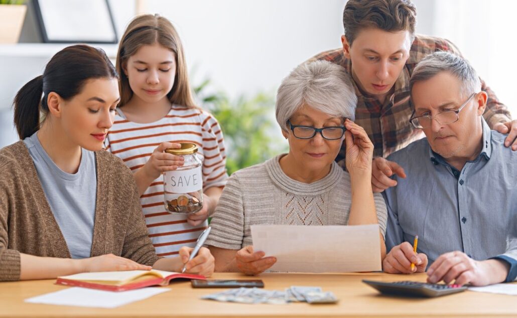Family members discussing financial planning at a table, with a senior woman reviewing documents, a young girl holding a savings jar, and two men analyzing expenses and calculations.